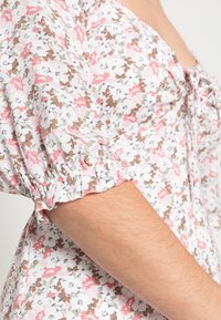 Blusa floral com padrões em rosa, branco e castanho, mangas bufantes com bordas franzidas e uma textura de tecido suave.