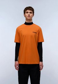 Oranje T-shirt met korte mouwen, gemaakt van katoen, met een klein bedrukt logo op de borst, gedragen over een zwart shirt met lange mouwen.