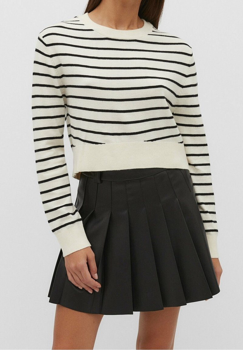Femme portant un pull blanc à manches longues avec des rayures horizontales noires et une mini-jupe plissée noire, debout les bras relâchés.