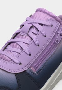 Κοντινή όψη μοβ αθλητικού παπουτσιού με ύφασμα από δίχτυ, διασταυρωμένα κορδόνια, ραμμένα επάνω στρώματα και πλαϊνό φερμουάρ σε λευκή σόλα.