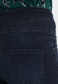 Vaqueros de mezclilla azul oscuro con cintura alta, que cuentan con un bolsillo trasero y detalles de costura sutiles. Textura suave.