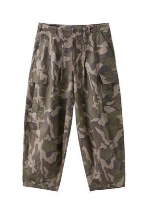 Ung kvinde med krøllet hår iført en hvid t-shirt med grøn kant og nummer 25 samt camouflage cargo bukser, stående mod en ensfarvet baggrund.