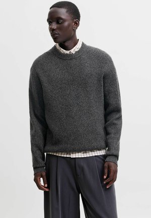 Personne portant un pull en laine gris foncé sur une chemise claire à carreaux et un pantalon ample foncé, debout devant un fond uni.