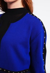 Pletený svetr s výrazným modrým tělem a černými akcenty, vysokým výstřihem a stříbrnými cvočky podél ramenního švu pro zvýraznění detailu.