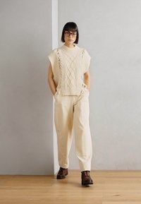 Top sin mangas de punto crema con patrones texturizados, combinado con pantalones beige y botas marrón oscuro, de pie frente a un fondo neutro.