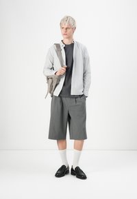 Grå korte bukser, sorte loafers, hvide strømper, sort t-shirt og en stribet skjorte med knapper. Bærer en lys grå crossover taske.
