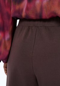 Bruine, rekbare stoffen broeken met een gladde textuur, voorzien van een hoge tailleband en zonder zichtbare hardware of patronen. Close-up van de achterkant.