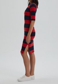 Donna che indossa un vestito a righe orizzontali rosso e blu navy, lungo fino al ginocchio, con uno spacco laterale e sneakers bianche, in posizione di profilo.
