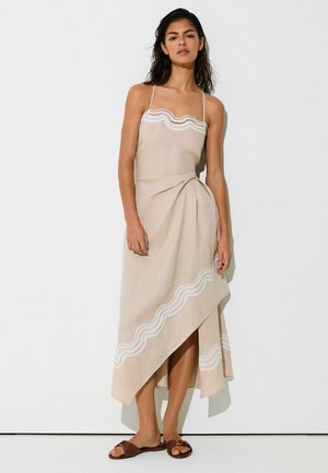 Kvinde iført en beige asymmetrisk ærmeløs kjole med hvidt blonderkant og brune flade sandaler, stående mod en hvid væg.