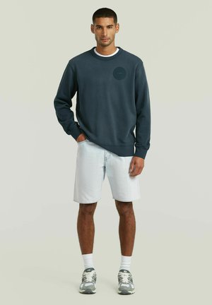 Homme debout en short en denim clair, sweat-shirt bleu foncé, chaussettes blanches et baskets grises sur un fond uni.