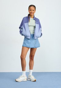 Giacca a vento color block viola e blu chiaro, top verde a scollo rotondo, minigonna di jeans, sneakers bianche con suola in gomma, su sfondo chiaro.