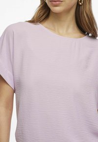 Gaiši lavandas krāsas īsām piedurknēm tops ar apaļo kakla izgriezumu un teksturētu audumu. Iespējas ietver brīvu piegriezumu un maigu kritumu.