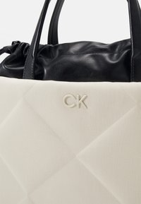 Sac fourre-tout beige et noir avec poignées et dessus en cuir souple, arborant le logo "CK" en relief sur le corps en tissu beige.