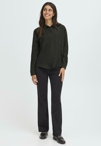 Woman standing, wearing black button-up shirt, black pants, and black loafers with gold chain detail, smiling and looking sideways.