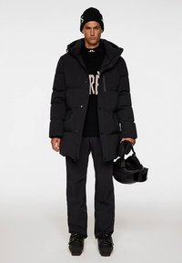 Crna prozračna jakna s kapuljačom, patentnim zatvaračem i kopčom na pritisak, nošena preko crnog džempera. Kombinirana s crnim hlačama i skijaškom kacigom u ruci.