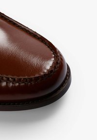 Zapato de cuero marrón con un acabado brillante, punta redondeada y costuras visibles a lo largo del borde. Textura suave y suela similar a madera.