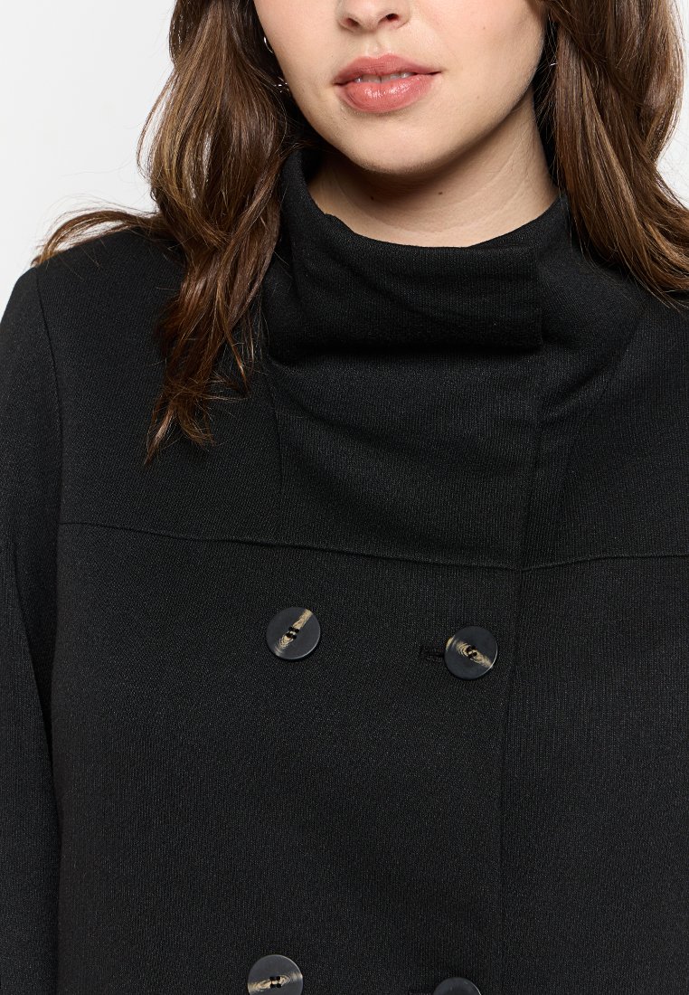 Cappotto doppio petto nero con colletto alto, caratterizzato da grandi bottoni rotondi e texture a coste, creando un aspetto strutturato ma morbido.