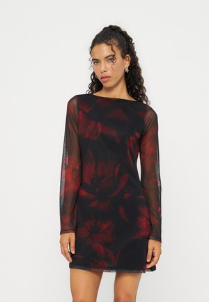 Femme aux cheveux bouclés portant une robe noire longue à manches transparentes avec des motifs floraux rouges, debout devant un fond clair uni.