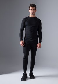 Top sportiv cu mânecă lungă, negru, cu decolteu în formă de cerc, asortat cu colanți negri, strânși și pantofi sport negri, pe un fundal gri neutru.
