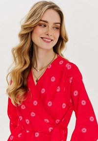 Červené zavinovací šaty s bílým květinovým potiskem, s véčkovým výstřihem, dlouhými rukávy a páskem v oblasti pasu, vyrobené z lehkého materiálu.