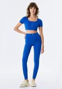 Top corto blu con maniche corte e zip frontale, abbinato a leggings blu a vita alta. Tessuto morbido ed elasticizzato con una superficie liscia.