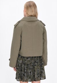Giacca cropped verde oliva con colletto, dettagli a bottoni e maniche raglan, abbinata a un vestito scuro a fiori. 