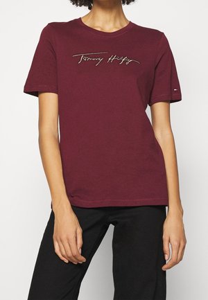 Persoon die een bordeauxrode korte mouw t-shirt draagt met de tekst "Tommy Hilfiger" op de borst en een zwarte broek tegen een witte achtergrond.