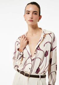 Romantisch crèmekleurig blouse met lange mouwen en een bordeauxrode bloemenpatroon, met een kraag, knoopsluiting aan de voorkant en een ontspannen pasvorm, gecombineerd met een bruine riem.