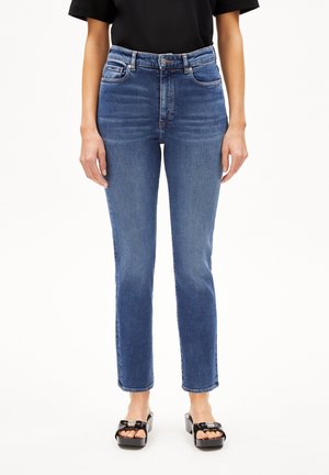 Femme portant un jean bleu droit taille haute, un haut noir à manches courtes et des sandales plateformes noires sur fond blanc.