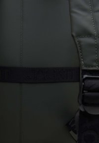 Svart rem med texten "JACKIT" på en mörkgrön texturerad tygbakgrund. Inkluderar sömdetaljer längs sömmen och spännet.