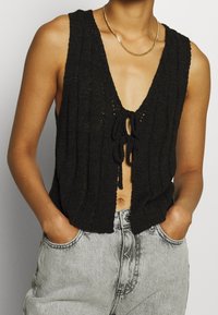 Black knitted vest with a ribbed texture, deep V-neck, and front tie closure, paired with high-waisted light gray denim shorts.