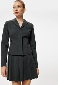 Traje de dos piezas en gris oscuro que cuenta con una chaqueta ajustada con solapas y una falda plisada. Diseño a medida con una textura de tela suave.