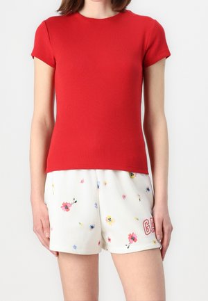 Vrouw draagt een rood T-shirt met korte mouwen en witte bloemenprintshorts met het "GAP"-logo op het linkerbeen, staand voor een effen achtergrond.