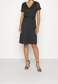 Černé šaty s krátkými průhlednými rukávy, výstřihem do V, opaskem na zavázání v pase, sukní ke kolenům, hladkou texturou, nošené s černými lodičkami na tenkých podpatcích.