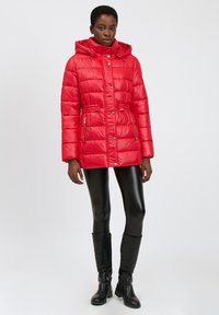 Chaqueta de plumón roja con capucha, bolsillos con cremallera y cintura ajustada. Textura suave, diseño acolchado, combinada con leggins negros y botas.