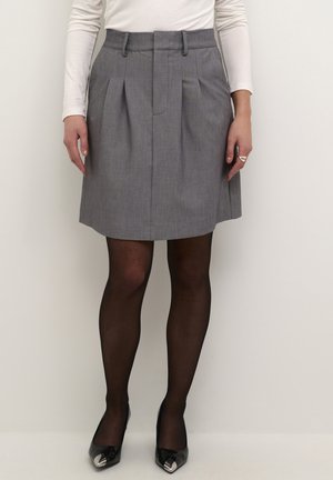 Femme portant une jupe plissée grise, des collants noirs transparents, des talons pointus noirs et un haut blanc à manches longues, debout devant un fond uni.