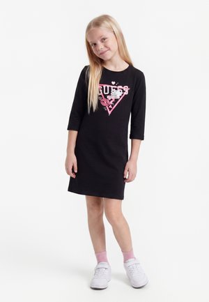 Ung pige med langt blondt hår iført en sort Guess-kjole, lyserøde sokker og hvide sneakers, stående med et let smil.