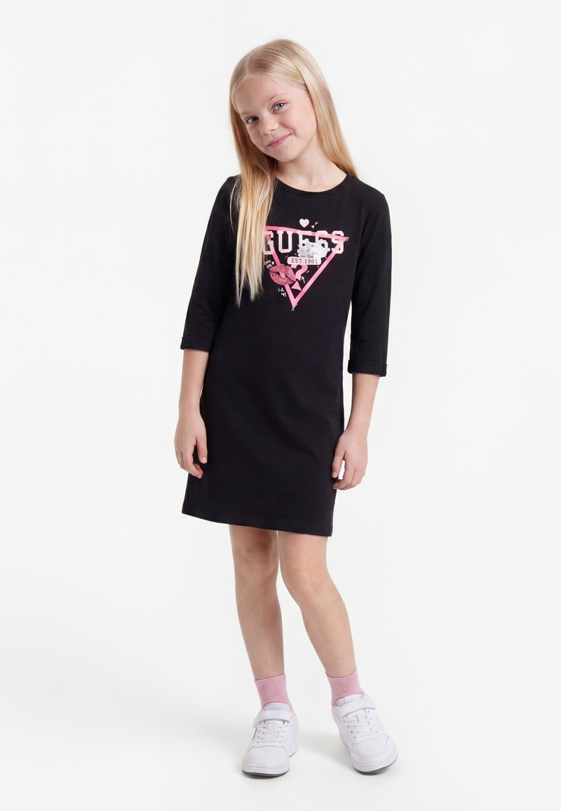 Jeune fille aux longs cheveux blonds portant une robe noire Guess, des chaussettes roses et des baskets blanches, debout avec un léger sourire.