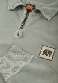 Sudadera de cremallera verde claro con una textura suave, puños acanalados y un logo de elefante marrón en un parche cuadrado.