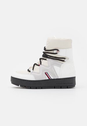 Botte d'hiver blanche et gris clair à tige haute, avec semelle épaisse noire, lacets noirs, et détails rouges, blancs et bleus sur le côté et aux œillets.