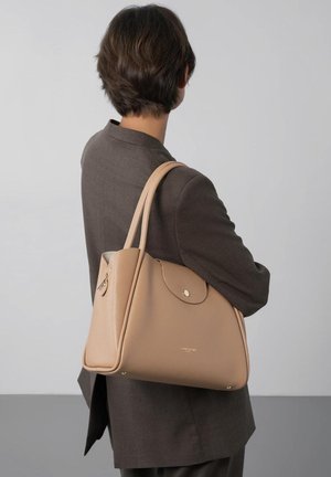 Personne aux cheveux courts et foncés portant un blazer sombre, avec un sac à main en cuir beige sur l'épaule, vue de dos sur un fond uni.