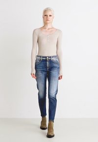 Béžový top s dlhými rukávmi a okrúhlym výstrihom, doplnený o detailné gombíky, v kombinácii so stredne umytými slim-fit džínsami a tanovými členkovými čižmami. Jednoduchý dizajn.