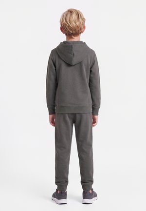 Barn med kort blondt hår iført en mørkegrå hættetrøje, matchende joggingbukser og grå sneakers, stående med ryggen mod på en hvid baggrund.