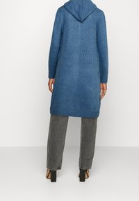 Lång, blå stickad cardigan med huva, texturerat mönster, ribbade ärmar och fåll, bärs över grå byxor och svarta klackar.