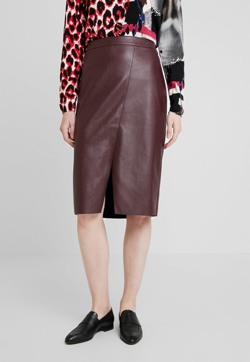 Femme portant une jupe en cuir marron foncé longueur genoux avec une fente devant, un chemisier à manches longues à motifs et des mocassins noirs, debout sur un sol uni.
