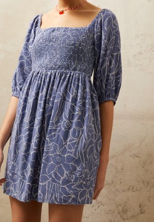 Robe bleue à motif floral blanc en contour, encolure carrée smockée, manches ballons trois-quarts, portée avec un collier de perles rouges orné d'un petit pendentif.