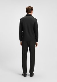 Veste noire ajustée avec un col doux, silhouette cintrée et manches longues. Associée à un pantalon noir et des chaussures sombres, vue de dos.