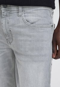 Nærbilde av lysegrå denimjeans båret av en person med mørk hud, som viser frontlomme, knapp og del av en svart genser.