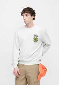 Hellgrauer Pullover mit langen Ärmeln, mit einem grünen, gestickten vierblättrigen Kleeblatt und Text. Kombiniert mit khakifarbenen Hosen und einer orangefarbenen Kappe.