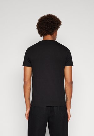 Hombre con cabello corto y rizado, vestido con una camiseta negra lisa de manga corta y pantalones negros, visto de espaldas sobre un fondo blanco.